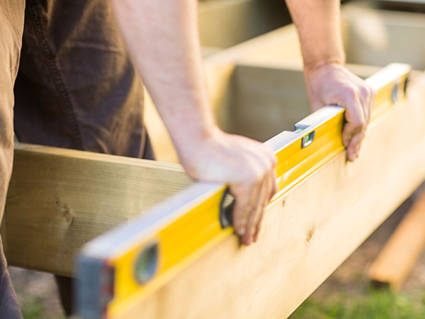 A close-up of two hands holding a yellow spirit level across a wooden beam, aligning it evenly during construction or carpentry work outdoors.