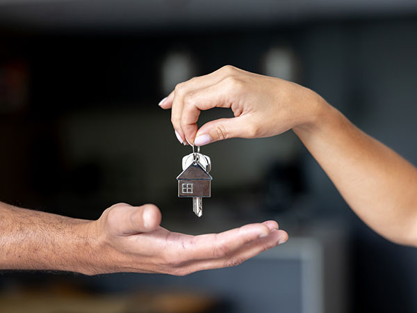 A close-up of one hand holding a keyring with a house-shaped keychain and keys above another open hand, suggesting the handover of house keys.