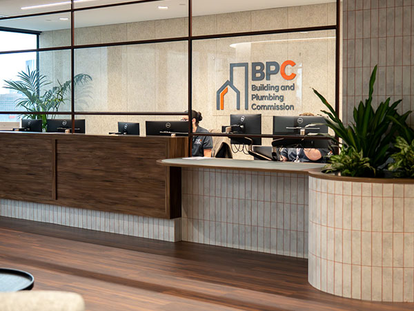 A modern reception area at the Building and Plumbing Commission (BPC) office, featuring a wooden front desk with computer stations and staff seated behind it, a glass wall displaying the BPC logo and name, and indoor plants positioned along the counter.