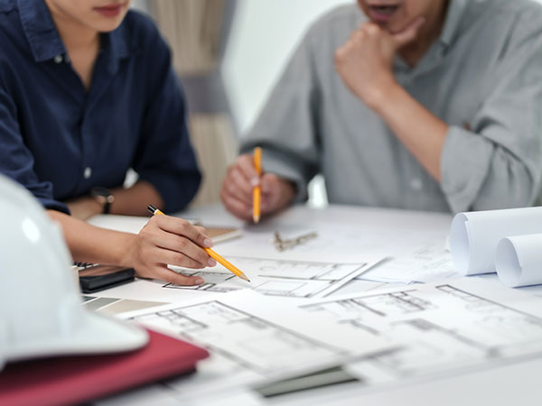 Two people sitting at a table reviewing architectural plans, pointing at drawings with pencils, with a laptop and samples visible, suggesting a design or planning discussion.