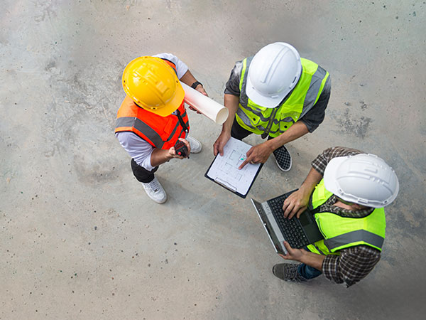 Overhead view of three construction workers wearing hard hats and high-visibility vests standing on a concrete surface, reviewing a plan on a clipboard while one uses a laptop and another holds rolled-up drawings.