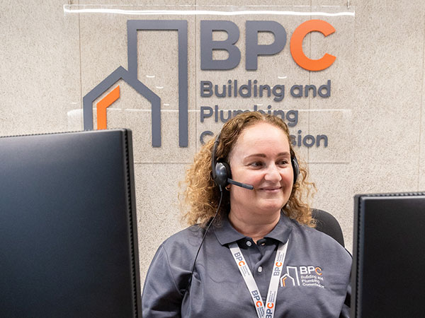 A BPC staff member wearing a headset and branded polo shirt sits between two computer monitors at a service desk, with a large Building and Plumbing Commission logo displayed on the wall behind.