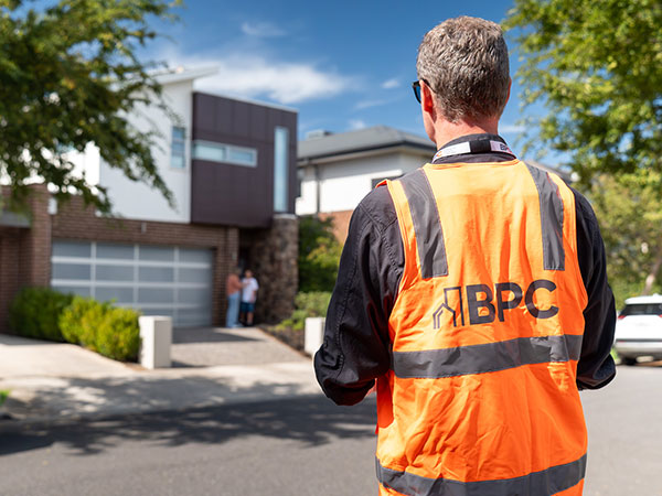 Inspector wearing an orange high-visibility vest marked “BPC” observing a modern residential house from the driveway.