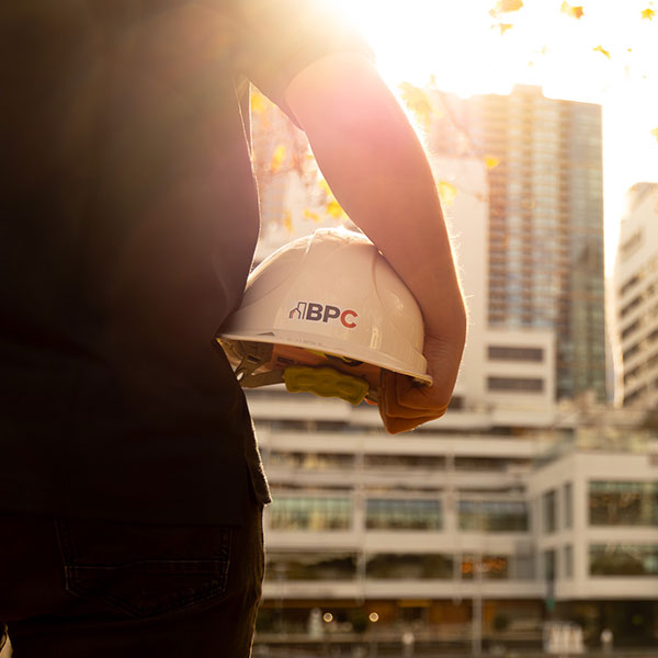 A close-up of a person holding a white hard hat with “BPC” printed on it, standing outdoors with city buildings in the background and warm sunlight shining from behind.