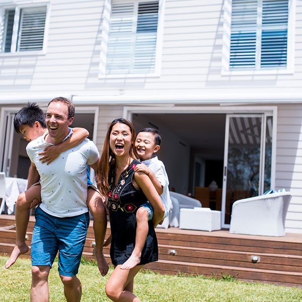A family playing outside in a backyard, with two adults each carrying a child on their back while walking across a lawn in front of a modern house with large sliding glass doors and a wooden deck.