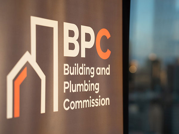 Close-up of BPC (Building and Plumbing Commission) signage on a glass wall, with the logo and text clearly visible and a blurred cityscape in the background.