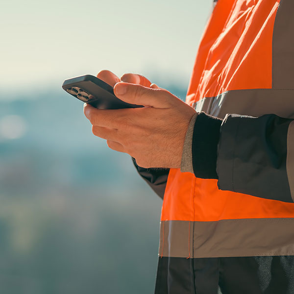 Close-up of a person wearing an orange high-visibility jacket holding and using a smartphone, with a blurred outdoor cityscape in the background.