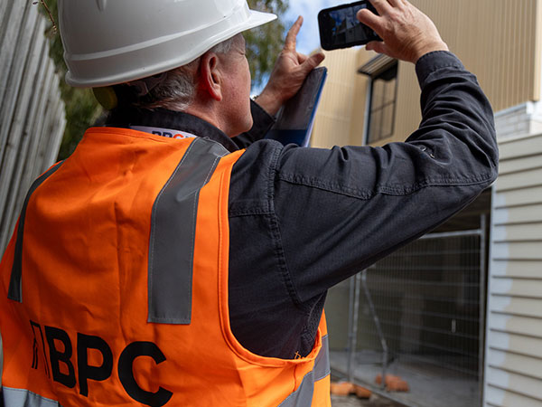 A close-up of a person wearing an orange high-visibility vest with “BPC” on the back and a white hard hat, holding up a smartphone to photograph the exterior of a building under construction.