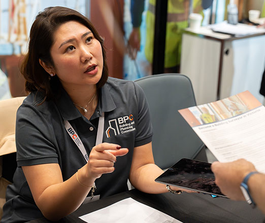 BPC staff member speaking with a visitor at an information booth, holding a tablet and discussing a printed brochure.