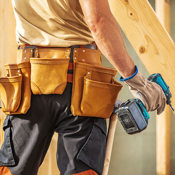A close-up of a person wearing a tool belt with multiple leather pouches, holding a cordless power drill while standing near wooden framing at a construction site.