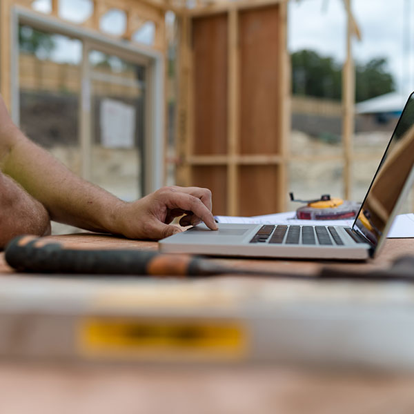 Close-up of a person’s hand using a laptop on a wooden workbench at a construction site, with timber framing of a partially built structure visible in the background and tools slightly blurred in the foreground.
