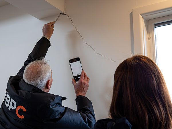 A close-up of two people inspecting a large crack running diagonally across an interior wall, with one person pointing at the damage while holding a smartphone and another observing, near a window.