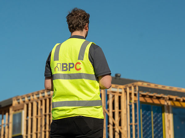 Rear view of a person wearing a high-visibility vest with “BPC” on the back, standing at a construction site and looking toward a partially framed timber house structure under a clear blue sky.