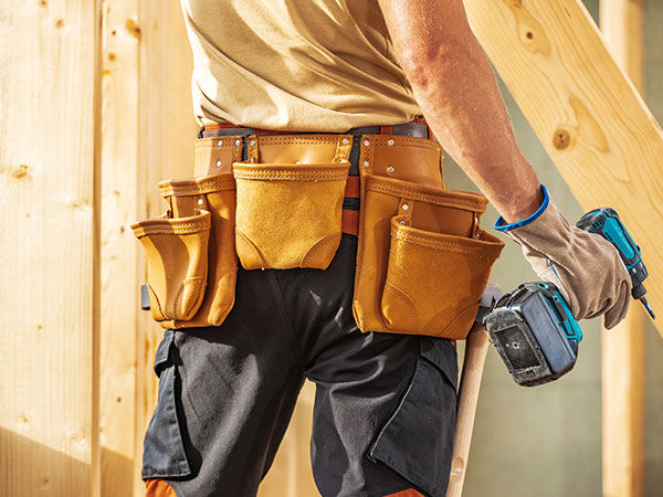 Close-up of a construction worker wearing a tool belt and holding a power drill next to a wooden frame.
