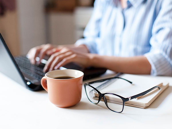Person typing on a laptop with a notebook, glasses, and a mug on a desk.