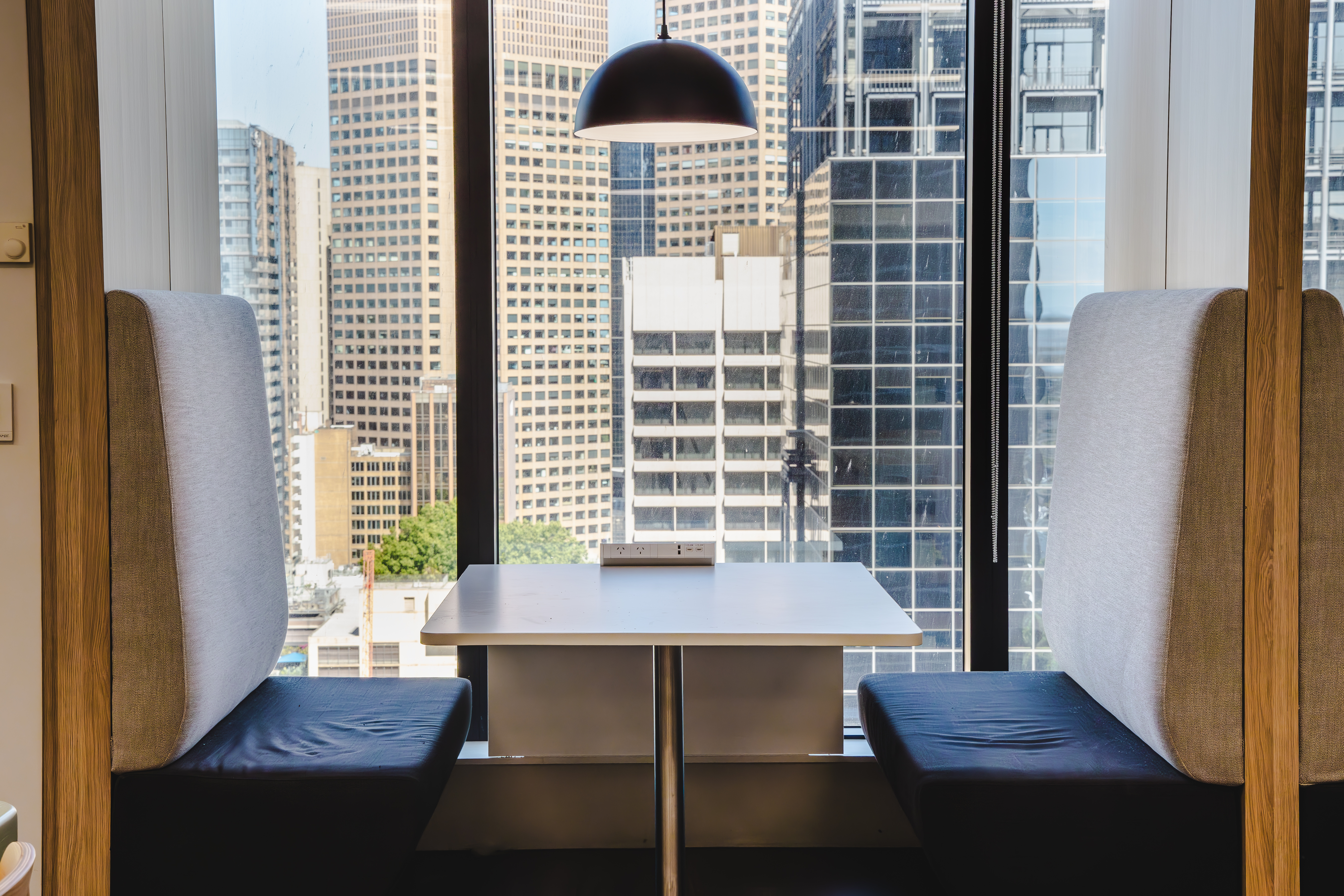 Quiet office booth with two cushioned bench seats facing a small table under a pendant light, beside a floor-to-ceiling window overlooking tall city buildings.