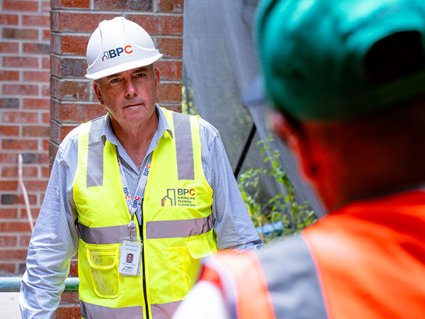 Person wearing a white hard hat and a high-visibility yellow vest with “BPC Building & Plumbing Commission” logo speaks to another worker in a high-visibility orange vest at a construction site, with a brick wall and equipment in the background.