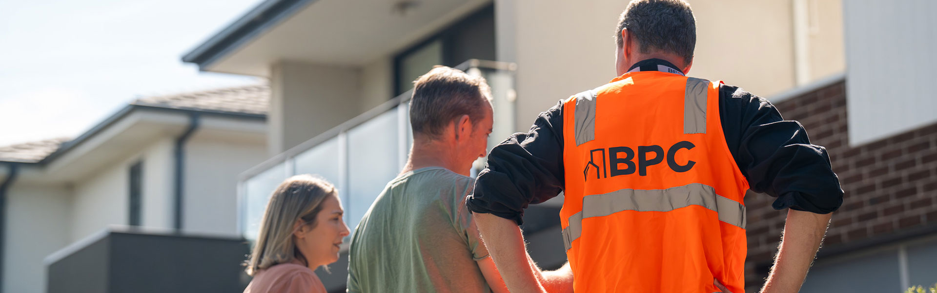 Person in a bright orange “BPC” hi-vis vest stands with two others outside a modern home.