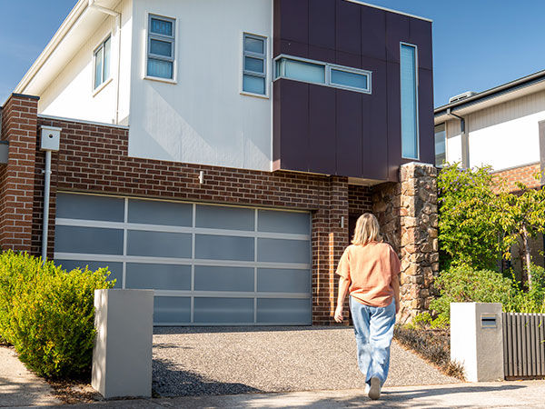 Person walking up a driveway toward a modern two-storey house with a double garage, featuring a brick and rendered exterior, large windows, and landscaped greenery.