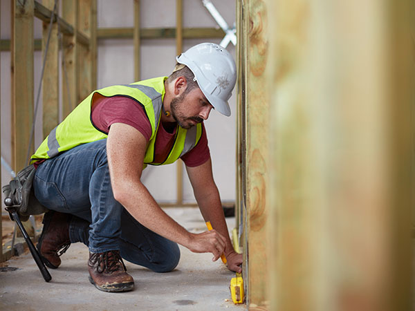 Construction worker in a hard hat and hi-vis vest kneels while measuring a wall frame indoors.
