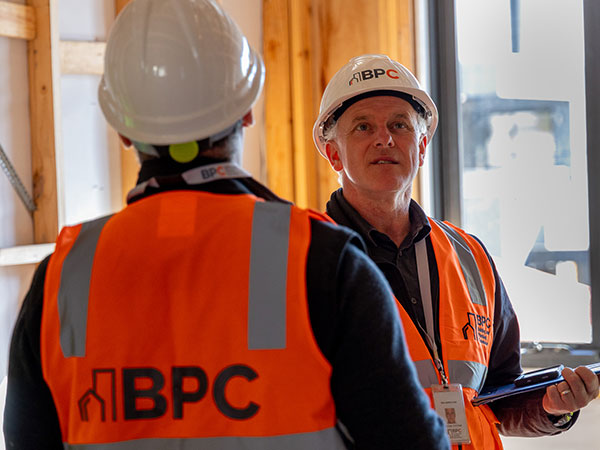 Two people in high-visibility vests and hard hats with “BPC” logos talk at a construction site, one holding a tablet.
