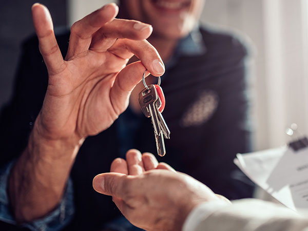 **Alt text:**   Close-up of one hand holding a set of house keys above another outstretched hand, indicating a handover, with a clipboard and paperwork partially visible in the background.